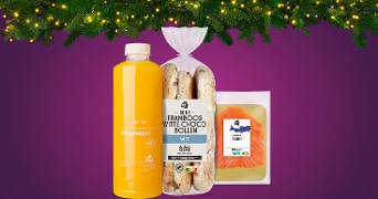 Fles vers sinaasappelsap, zakje mini framboos witte choco bollen en verpakking gerookte zalm van Albert Heijn op een paarse achtergrond met kerstverlichting erboven