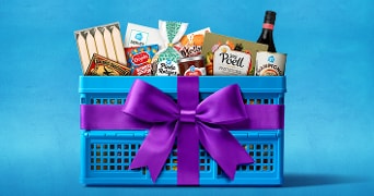 Blauwe krat met paarse strik gevuld met Albert Heijn huismerkproducten, snacks, servetten, een fles wijn en Jos Poell pasteitjes tegen een blauwe achtergrond