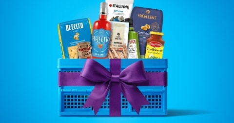 Blauwe geschenkmand met Italiaanse etenswaren zoals De Cecco pasta, Perfeito saus, Grand'Italia pastasaus, Excellent conchiglie en diverse andere producten, versierd met een paarse strik.