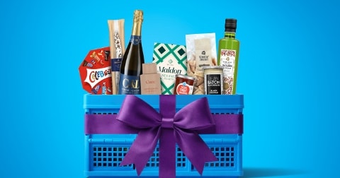 Blauwe krat gevuld met diverse A-merk producten, waaronder champagne, olijfolie, crackers, zout, en koekjes, versierd met een paarse strik tegen een blauwe achtergrond.