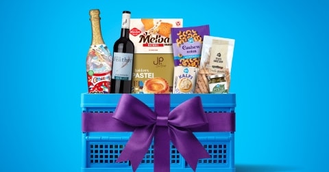 Blauwe Albert Heijn krat gevuld met een selectie feestelijke producten, waaronder een fles wijn, bubbeldrank, Melba toast, bakker's pastéi, cashewnootjes en andere snacks, versierd met een paarse strik tegen een blauwe achtergrond.