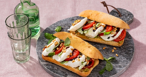 Ciabatta broodjes met mozzarella, tomaat, basilicum en pijnboompitten op een stenen bord, naast een groene glazen fles en glazen.