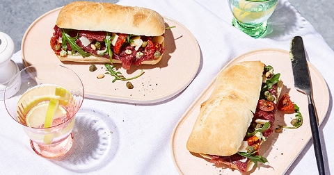 Twee ciabatta broodjes met rucola, gedroogde tomaten en kappertjes geserveerd naast een glas drank met een citroenschijfje.