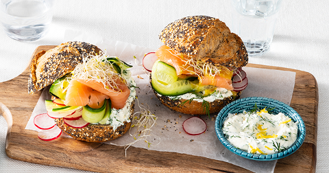 Volkorenbroodjes met gerookte zalm, roomkaas, komkommer, radijs en alfalfa op een houten plank, met een klein schaaltje kruidenroomkaas.