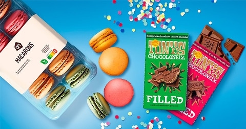 Macarons van Albert Heijn in verschillende kleuren naast twee repen Tony's Chocolonely chocolade, met gekleurde confetti op een blauwe achtergrond.