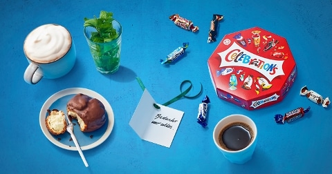 Ontbijtscène met Celebrations chocolaatjes, koffie, een mok met slagroom, een glas groene kruiden en een bord met een gebakje.