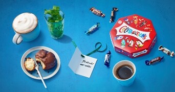 Ontbijtscène met Celebrations chocolaatjes, koffie, een mok met slagroom, een glas groene kruiden en een bord met een gebakje.