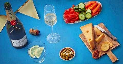 Fles Moët & Chandon champagne met hapjes zoals kaas, gesneden paprika, komkommer, wortel, olijven en citroenschijfjes op een blauwe achtergrond.