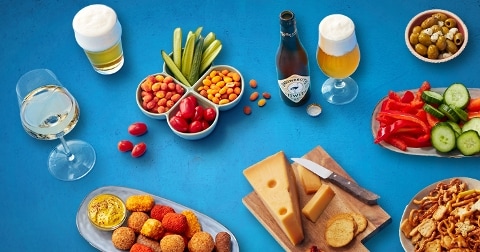 Borrelplank met kaas, snacks, groenten, olijven en drankjes op een blauwe achtergrond.