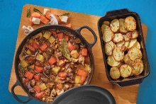 Stoofpot met rundvlees, wortel, aardappel en champignons in een pan naast een ovenschaal met gebakken aardappelschijfjes, geserveerd op een houten plank
