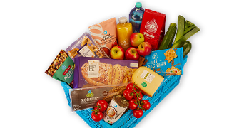 Blauwe boodschappenmand gevuld met diverse Albert Heijn producten zoals appels, cherrytomaten, komkommers, kaas, mini crackers, feeststol, fles water, vruchtensap, koffie van Perla en AH notenmix.
