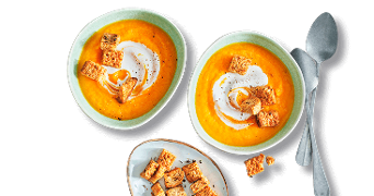 Twee kommen pompoensoep met room en croutons, geserveerd met lepels en een schaaltje extra croutons