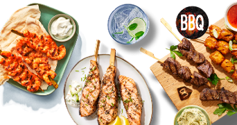 Verschillende BBQ-spiesen met gegrilde garnalen, zalm en rundvlees, geserveerd met sausjes en een glas frisdrank met munt en limoen.