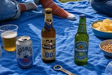 Drie soorten alcoholvrij bier, waaronder Bud, Hertog Jan en Heineken, staan op een blauw picknickkleed met snacks en een biertje in een plastic beker.