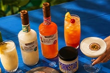 Twee flessen ready-to-serve cocktails met diverse zomerse cocktails in glazen op een blauwe tafel, waaronder Piña Colada en Pornstar Martini.