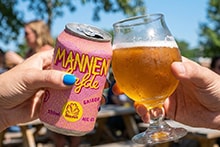 Twee mensen proosten met een blik Mannen Liefde bier en een gevuld bierglas buiten op een zonnig terras.