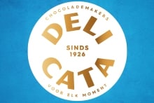 Logo van Delicata chocolademakers, met goudkleurige tekst op een witte cirkel tegen een blauwe achtergrond.