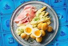 Bord met witte asperges, ham, krieltjes en een gehalveerd gekookt ei, gegarneerd met bieslook, op een blauw tegeltjesservet.