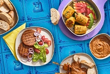Gegrilde hamburgers met sla, tomaat, ui en brood, geroosterde groenten zoals maïs en paprika, saté met pindasaus en brood, gepresenteerd op borden tegen een blauwe achtergrond.