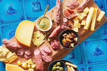 Borrelplank met verschillende kazen, worst, noten en olijven op een blauwe achtergrond.