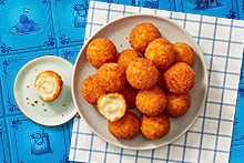 Bord met gefrituurde kaasballetjes, waarvan één opengebroken met gesmolten kaasvulling, op een geruite theedoek.