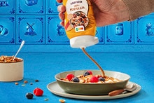 Pindakaas wordt over een ontbijtkom met yoghurt, granola, aardbeien en blauwe bessen gegoten, met een potje granola op de achtergrond.