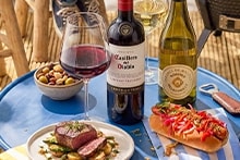 Tafel met een bord met vlees en groenten, een hotdog, een kom met gemengde noten, een glas rode wijn, een glas witte wijn, een fles Casillero del Diablo rode wijn en een fles witte wijn.