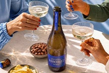 Fles Lahoré sparkling wijn op een tafel met drie mensen die proosten met glazen, omringd door borrelhapjes zoals chips en noten.