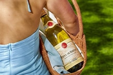 Fles witte wijn Côtes du Rhône in een rieten tas, gedragen door een persoon in zomerse kleding op een grasveld