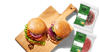 Twee gepofte hamburgerbroodjes met verse toppings op een houten serveerplank, naast twee verpakkingen Greenfields rundergehakt van Albert Heijn.