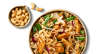 Roerbakgerecht met noedels, kip, cashewnoten en groenten in een kom, met een apart bakje cashewnoten.