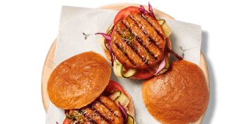 Twee hamburgers op een houten plank, bekleed met bakpapier, gevuld met gegrilde burger, tomaat, komkommer en rode ui.