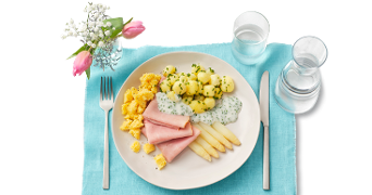 Bord met ham, witte asperges, krieltjes met kruiden, hollandaisesaus en roerei op een blauw placemat, met bestek, glas water, karaf en een vaasje bloemen.