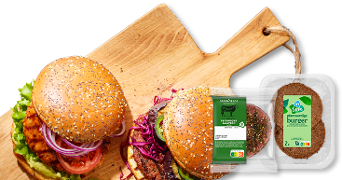 Twee broodjes hamburger op een houten snijplank, belegd met verse groenten, naast verpakkingen van AH plantaardige burger en Greenfields belegen kaas.
