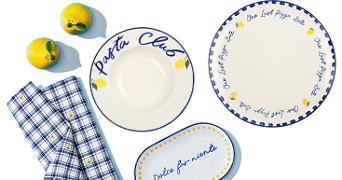 Wit servies met blauwe en gele details, waaronder een diep bord met de tekst "Pasta Club", een rond bord met de tekst "One Last Pizza Bite", een ovale schaal met de tekst "Dolce far niente", twee blauwe geruite servetten en twee citroenen.