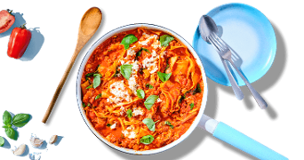 Lasagne in een pan met tomatensaus, basilicum en kaas, omringd door een houten lepel, een blauw bord met bestek en verse ingrediënten zoals tomaat, basilicum en knoflook.