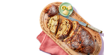 Gesneden stolbrood met rozijnen en amandelspijs op een houten plank, met een schaaltje boter en een mes.