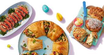 Verschillende feestelijke Paasgerechten op tafel, waaronder een salade met zalm en sesamzaadjes, een hartige ronde plaattaart gevuld met groenten, amandelbrood en zoete broodjes met kokos, omringd door decoratieve paaseitjes.