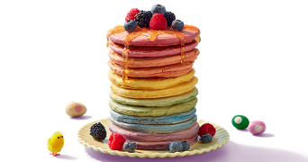 Stapel kleurrijke pannenkoeken met honing en vers fruit zoals bramen, frambozen en blauwe bessen, geserveerd op een bord met paassnoepjes en decoratief paaskuikentje.