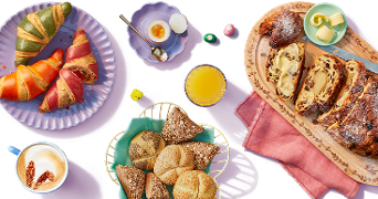 Ontbijttafel met kleurrijke croissants, verschillende soorten broodjes met zaden, plakjes gevulde kerststol op een houten plank met roomboter, een zachtgekookt ei, een glas sinaasappelsap en een kop cappuccino.