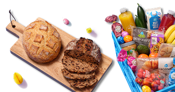 Vers brood op een houten plank naast een blauwe Albert Heijn boodschappentas gevuld met diverse boodschappen zoals fruit, groente, zuivel, vruchtensappen, croissants en andere verpakte producten.