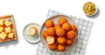 Gefrituurde kaasballetjes op een bord, geserveerd met blokjes kaas en een schaaltje mosterd op een tafel met een geruit servet.
