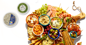 Borrelplank met verschillende hapjes waaronder druiven, olijven, toast, gedroogde worst, kaas, geroosterde groenten, garnalen en diverse dips zoals AH Excellent tonijnsalade en groene pesto.