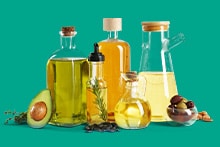 Verschillende soorten plantaardige olie in glazen flessen, omringd door avocado en olijven, tegen een groene achtergrond