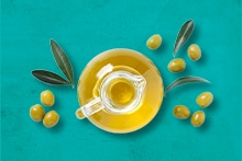 Glazen kan met olijfolie omringd door verse olijven en olijftakken på een turquoise achtergrond.