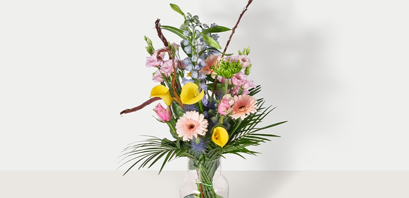 Boeket met diverse kleurrijke bloemen, waaronder gele calla lelies, roze gerbera’s en groene bladeren in een glazen vaas