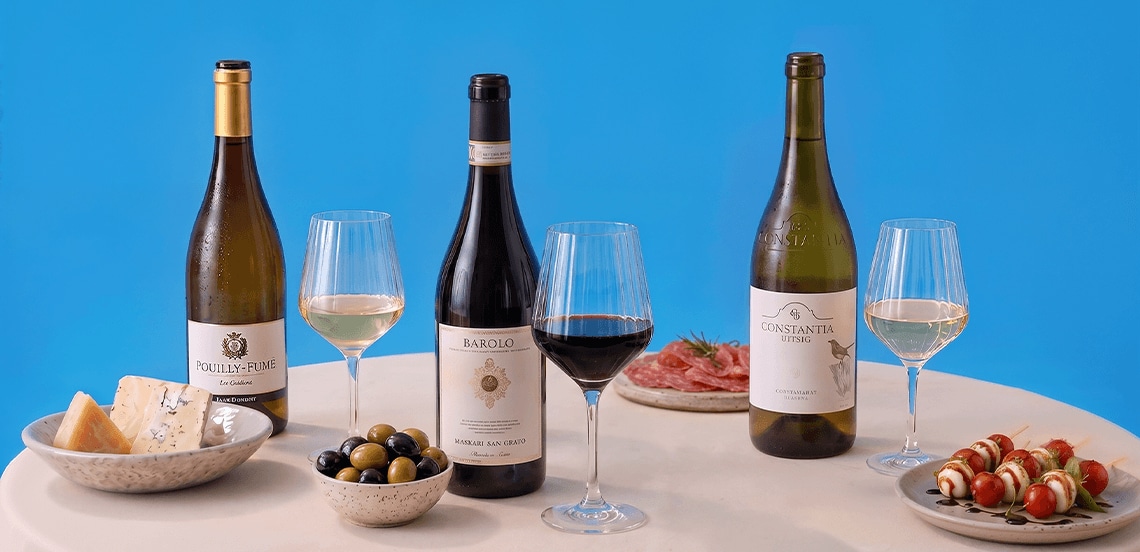 Drie flessen wijn (Pouilly-Fumé, Barolo, Constantia Uitsig) met drie bijpassende wijnglazen op een tafel, geserveerd met een kaasplankje, een schaaltje olijven, een bordje vleeswaren en een bordje spiesjes met mozzarella en tomaat.