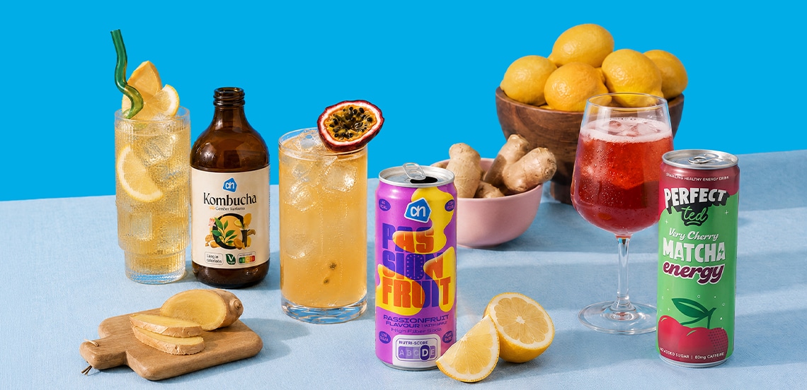 Assortiment van verfrissende drankjes, waaronder Albert Heijn Kombucha, passievrucht frisdrank en Perfect Ted Matcha Energy, gepresenteerd met glazen, ijs, schijfjes citroen, gember en een schaal met citroenen en gember op een blauwe achtergrond.