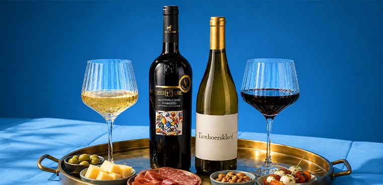 Twee glazen wijn met flessen rode en witte wijn van de merken Montepulciano d’Abruzzo en Tamboerskloof op een gouden serveerschaal, geserveerd met kaas, olijven, worst, noten en hapjes.