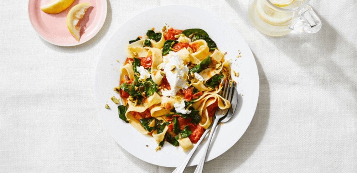 Tagliatelle met spinazie, tomaat en ricotta op een wit bord, geserveerd met citroenpartjes.